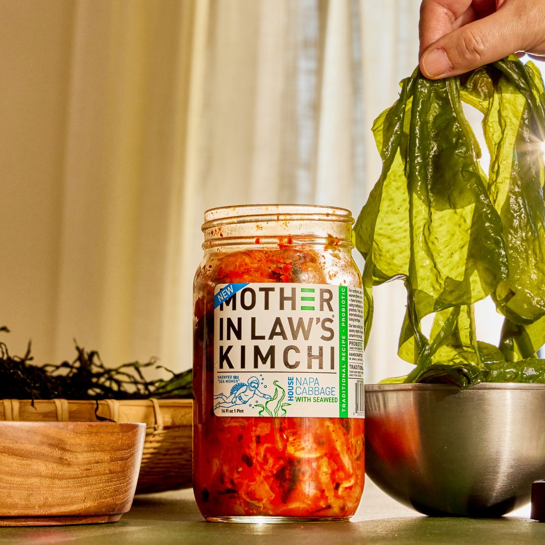 A jar of Mother-in-Law’s kimchi on a counter beside fresh seaweed being lifted from a bowl